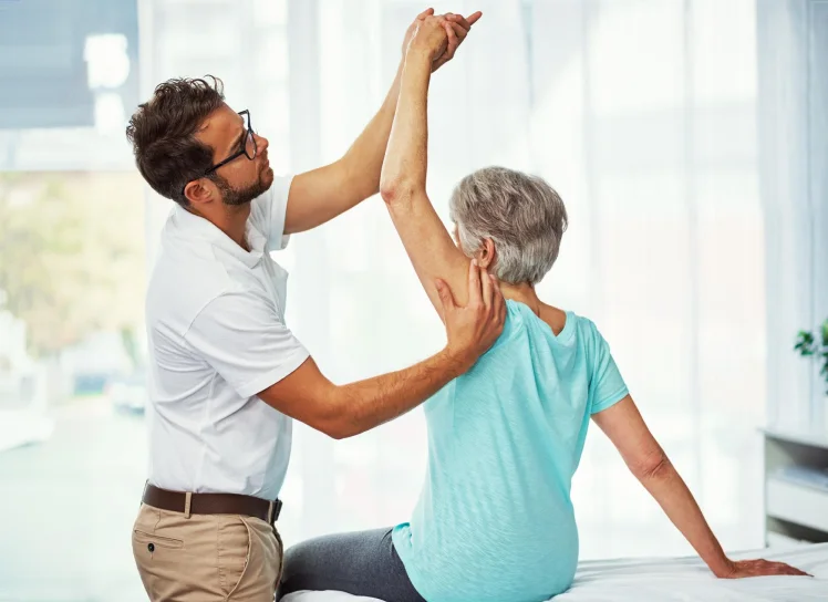 Therapist guiding an older woman through a shoulder exercise at home.