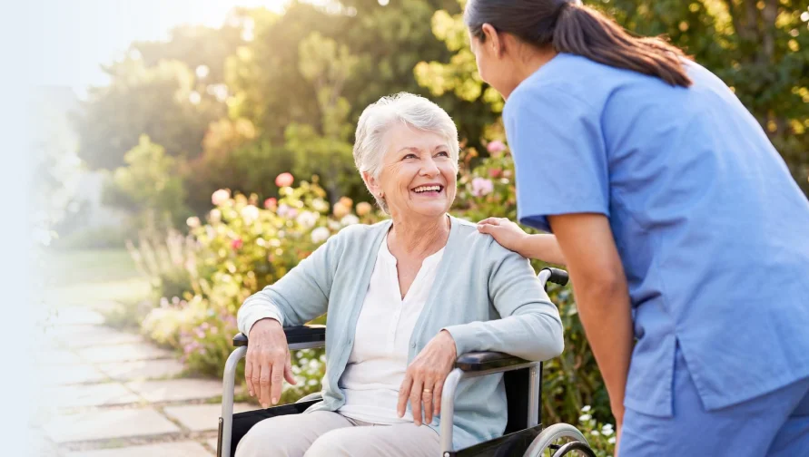 Caregiver speaking with an older woman in a wheelchair outdoors.