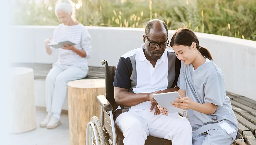 Caregiver showing a tablet to a senior man using a wheelchair outdoors